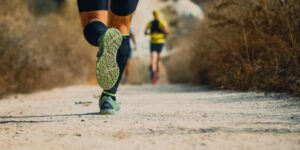 Dos personas corriendo por un camino una delante y otra detrás. A la persona que está delante solo se ven las piernas y las zapatillas.