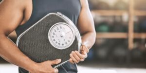 Hombre con una camiseta de tirantes llevando una báscula entre sus brazos.