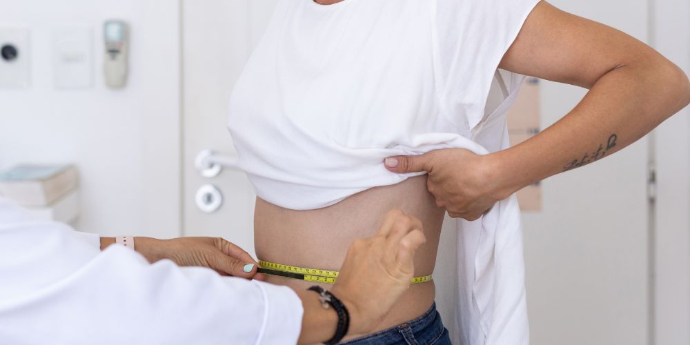 Médico midiendo la cintura de una mujer con la camiseta levantada con una cinta de tomar medidas.
