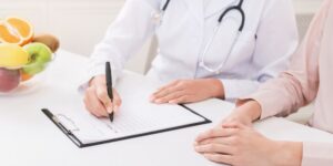 Un médico con un fonendoscopio escribiendo a un paciente que está a su derecha con un bolígrafo y un papel.