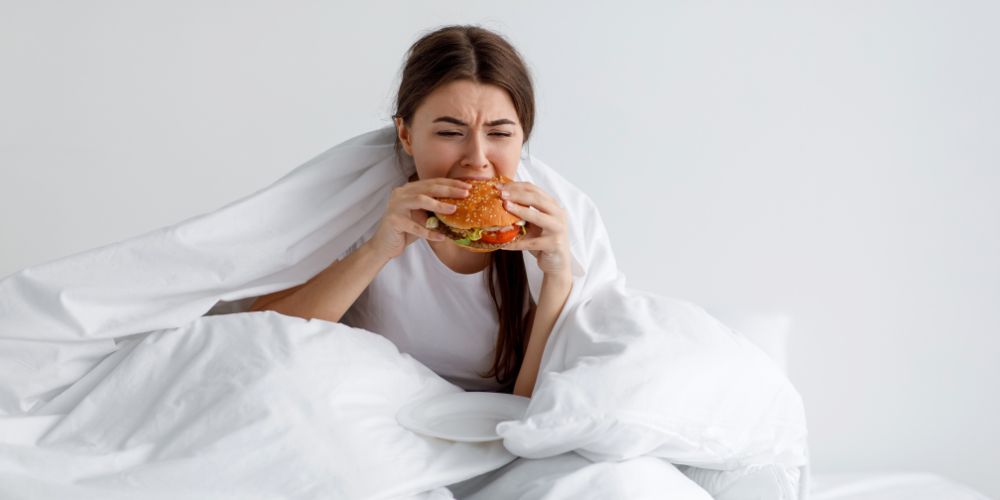Mujer comiendo una hamburguesa con ansiedad en la cama.