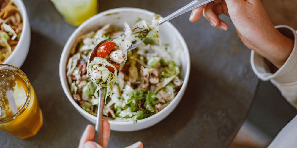 Un plato con una ensalada y dos manos con un cuchillo y tenedor cogiendola.