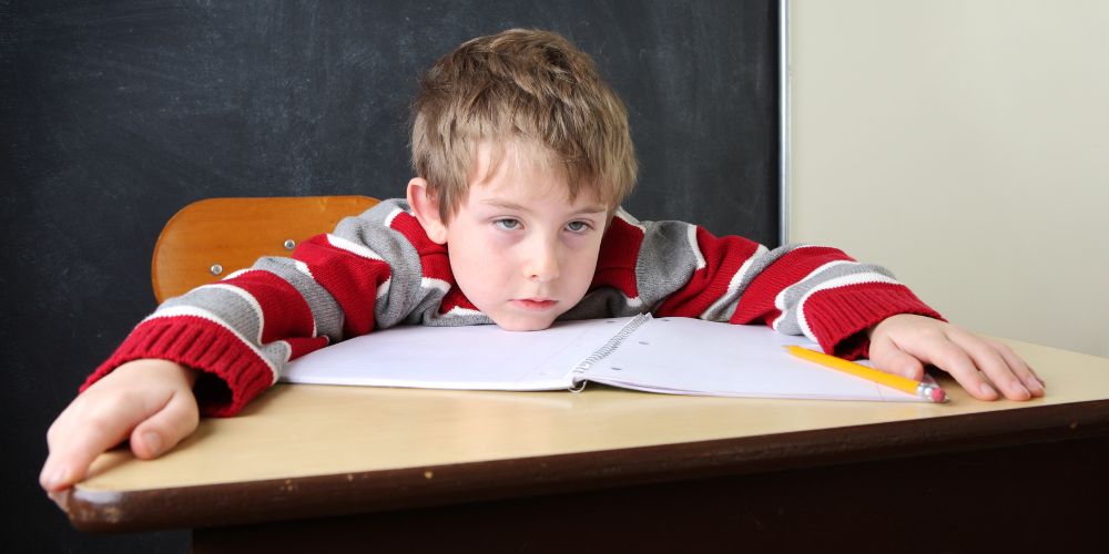 Niño apoyado en la mesa con los brazos abiertos. Debajo de su cara tiene una libreta abierta y un lápiz.