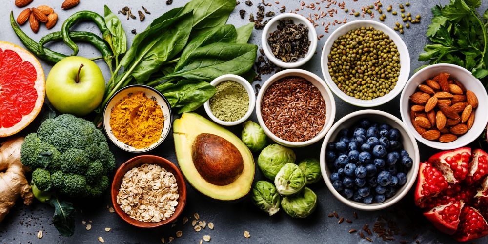 Frutas y verduras en una mesa: aguacates, arándanos, una manzana, broccoli, una granada, etc...