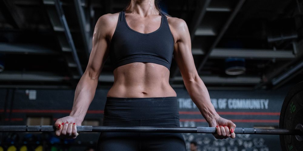 Mujer levantando peso en un gimnasio con los brazos extendidos y el abdomen al aire.