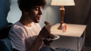 hombre en un sofá mirando una galleta que cree que no debe comerse, al fondo una lámpara y un reloj